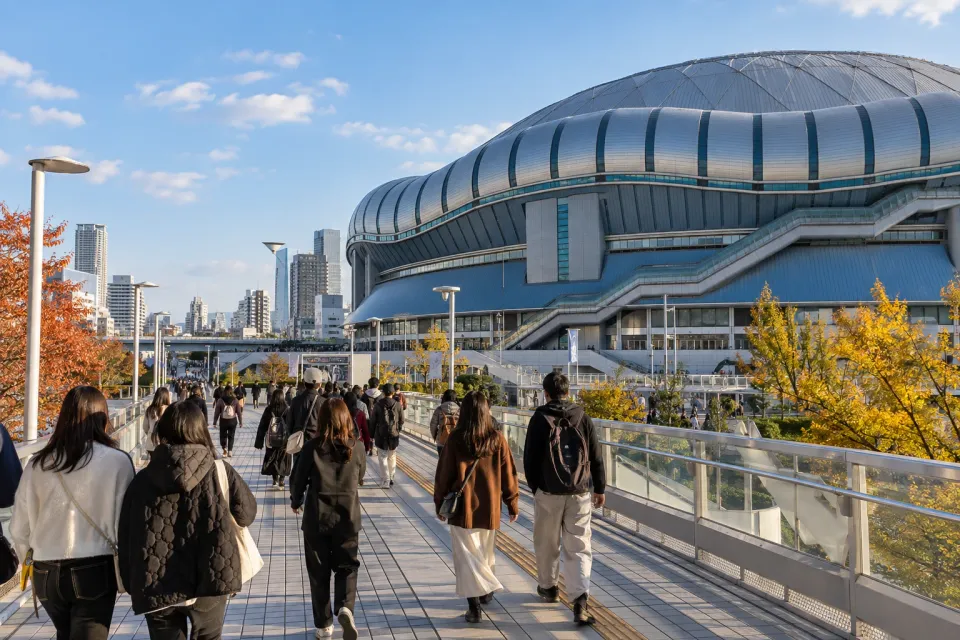 Generated travel editorial image of the pedestrian approach to Kyocera Dome Osaka in autumn